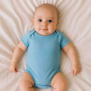 A toddler wearing a light blue baby bodysuit while lying on a soft cotton bedsheet