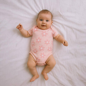 A toddler is wearing a floral pink cutie baby bodysuit while lying on a soft cotton bedsheet
